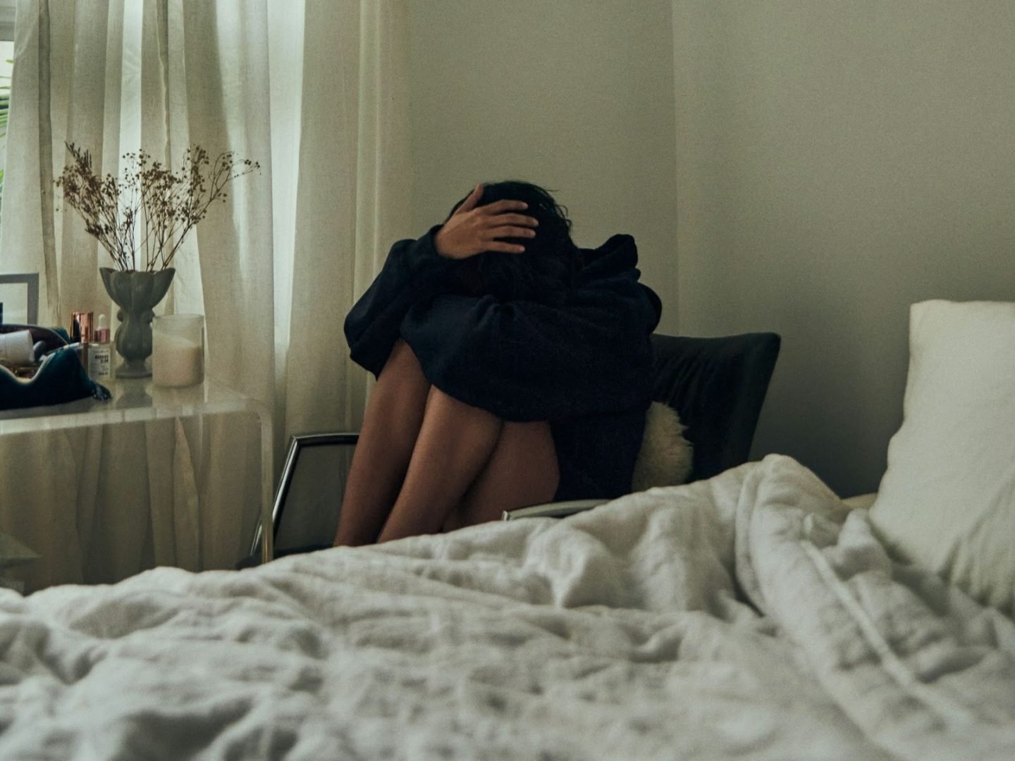 Person sitting curled up on a chair beside a bed, head down and arms wrapped around knees, in a softly lit bedroom with curtains and minimal decor, conveying emotional distress, isolation, or overwhelm.