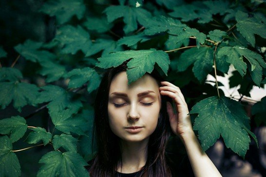 Woman with closed eyes standing beneath green leaves in a calm and introspective moment in nature.