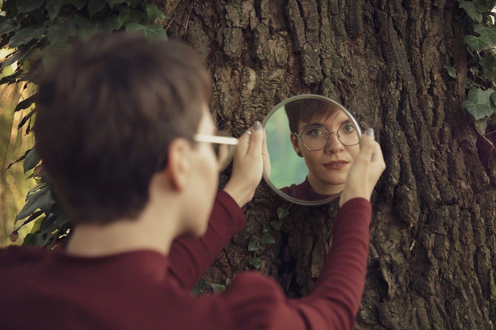 Person holding a mirror against a tree while looking at their reflection in a peaceful outdoor setting.